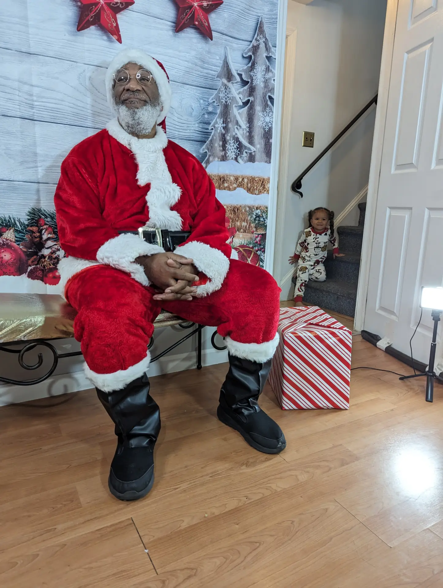 Authentic Black Santa Bruce sitting on a bench in a festive holiday setting, with a toddler in Christmas pajamas peeking from the staircase in the background. The photo conveys the anticipation and magic of meeting a diverse Santa Claus.