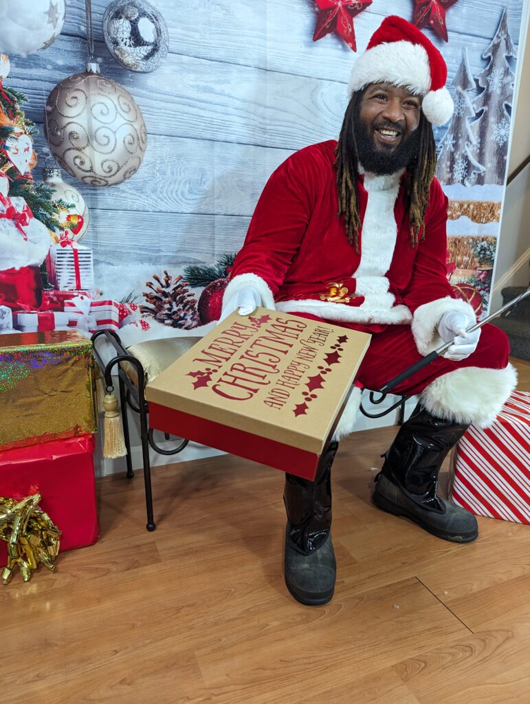 Santa Chris, an authentic Black Santa Claus with dreadlocks, sitting in a festive holiday backdrop, holding a gift box. He is smiling and ready to spread cheer, representing the joyful spirit of Selfies with Santa appearances for events and photo shoots.