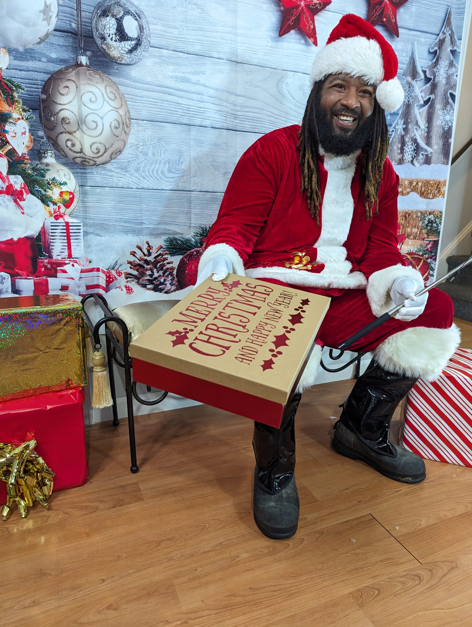 Santa Chris, an authentic Black Santa Claus with dreadlocks, sitting in a festive holiday backdrop, holding a gift box. He is smiling and ready to spread cheer, representing the joyful spirit of Selfies with Santa appearances for events and photo shoots.